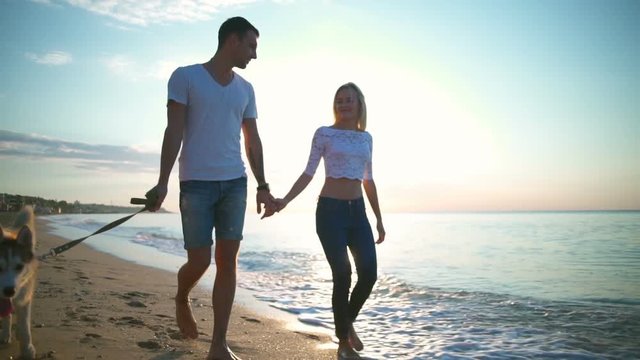 Young pretty couple smiling walking with dog at seaside at sunrise. Slow motion.