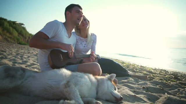 Couple smiling sitting with dog near sea. Man playing ukulele. Slow motion.