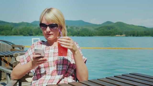 Woman resting on a summer area in the cafe. It uses the phone. In the background, the lake and the mountains. Resort in Spain