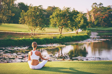 Yoga at park with view of the mountains, with sunlight. Young woman in lotus pose sitting on green grass. Concept of calm and meditation.