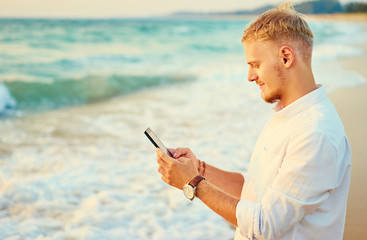 Vacation and technology. Portrait of young handsome man using smartphone on the sea shore.