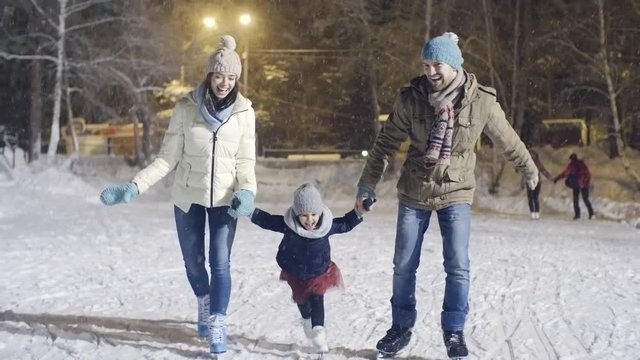 Dolly shot of laughing young woman and bearded man holding hands of little girl and skating on ice rink on snowy winter evening