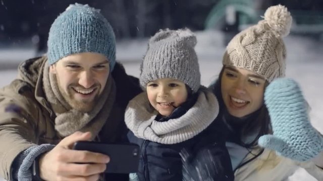 Panning shot of young family laughing and video calling someone in park on snowy winter evening