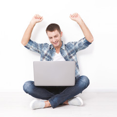 Handsome young man sitting on the floor near the wall, celebrating success with arms raised while looking at his laptop screen