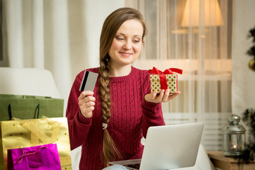 Happy young woman using laptop sitting in the living room with decorated Christmas tree. Bright shopping bags, Candles and lights. Holding in hands credit card. E-commerce and online shopping concept