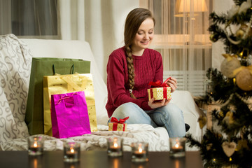 Obraz na płótnie Canvas Happy young woman in red sweater sitting in the living room with decorated Christmas tree. Bright shopping bags, Candles and lights. Opening golden gift box with red ribbon. Happy holiday concept. 
