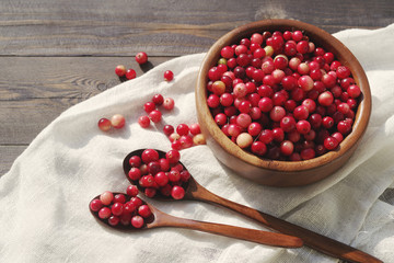 Fresh red forest cranberry in a round bowl with a wooden spoon on white linen fabric on a table surface. Autumn harvest of wild berries