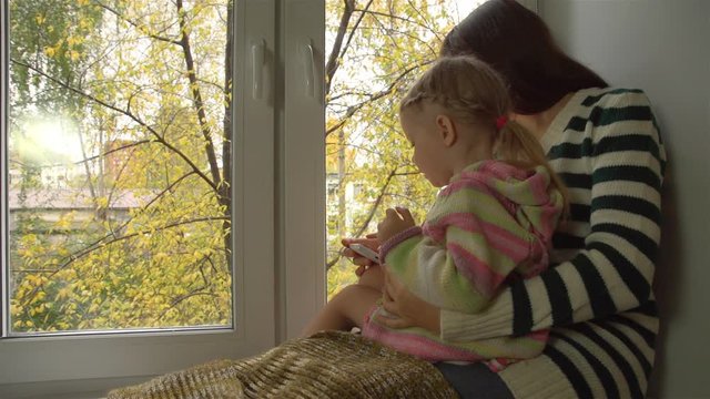 Autumn day, mother and daughter playing on the windowsill in the smart phone.