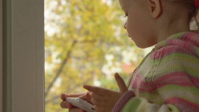 Маленькая девочка в полосатой кофте играет в игру на смартфоне, сидят на подоконнике. Смартфон поддерживает женская рука. За окном желтые листья на деревьях.