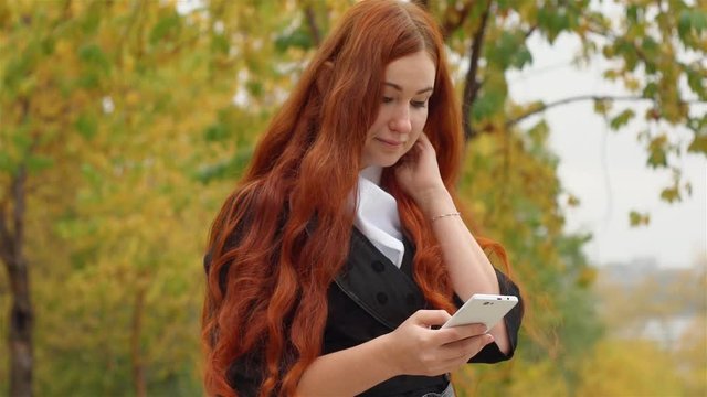 Girl in black coat  red long hair walking in autumn park, talking with friends on the social networks on the touch phone her good mood. Yellow leaves falling from the trees.