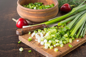  Green onions.   Green onions on a cutting board and in a wooden bowl, knife and pods of red hot pepper on old brown table.