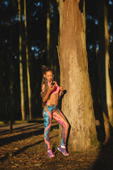 Sporty black beautiful woman taking a fitness workout rest for playing music or texting on her smartphone. Brazilian girl in colorful fashion sportswear training in nature under sunrise light.