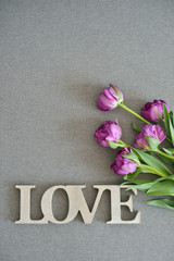 Fresh spring purple tulips flowers bouquet on grey canvas background, word love with wooden texture. Place for text. Green tulips petals, tulips and wood.