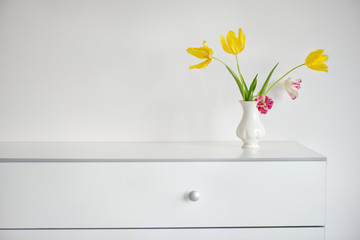 interior design scene with a white modern commode and a vase of yellow and pink tulips on a pale...