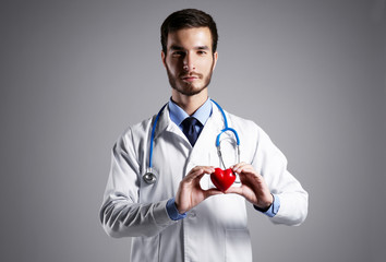 Handsome male doctor holding red heart on grey background