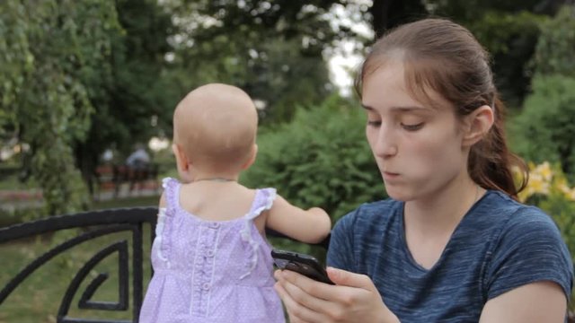 A young nanny plunging into a picture on her smartphone s screen and holding a baby girl with one hand.