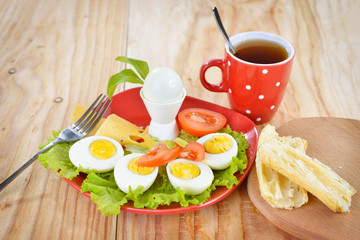 Breakfast with hard boiled eggs, sliced in halves, cup of tea, salad, tomatoes, cheese and bread on the red plate and wooden background