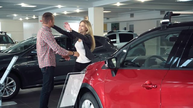 Man Standing Behind Woman and Covering Her Eyes While Standing in front of Shiny New Red Vehicle Inside Car Dealership - Man Surprising Woman with New Car in Show Room.