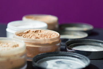Mineral make up powder for matt skin. Variety of containers with beige and white powder for facial care. Focus on beige powder, purple background