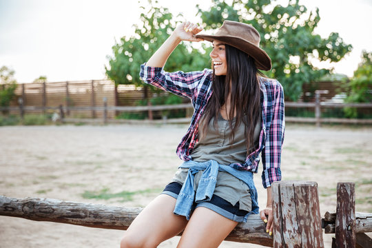 Cheerful relaxed young woman cowgirl sitting on fence and smiling