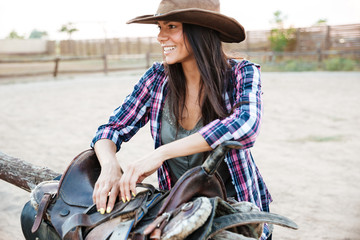 Cheerful young woman cowgirl preparing saddle for riding horse