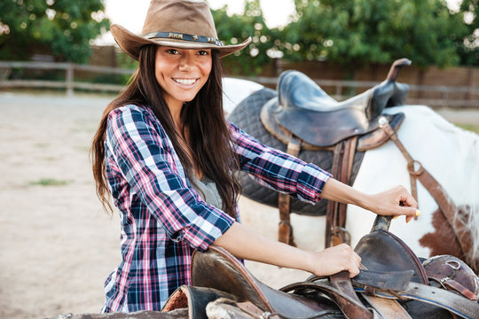 Smiling woman cowgirl standing and preparing saddle for riding horse