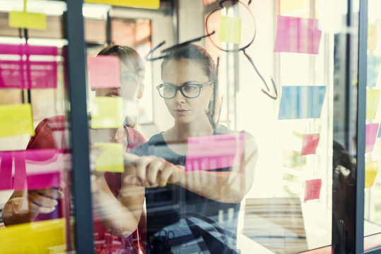 Two business women working together on wall glass with post-it 