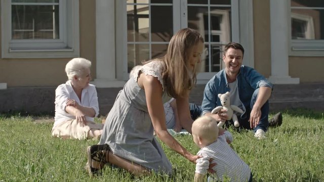 Blond toddler boy surrounded by supporting and loving family learning to walk but constantly falling on green grass