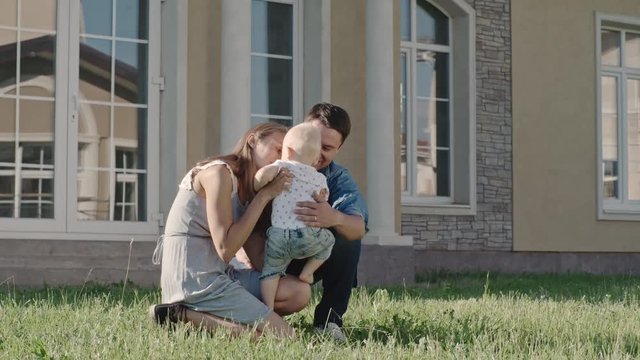 Lockdown of loving parents sitting on green grass in backyard hugging and kissing their blond toddler boy