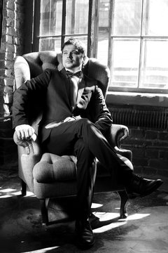 Handsome man in black formal suit with bow-tie sitting in armchair. Black and white fashion style portrait