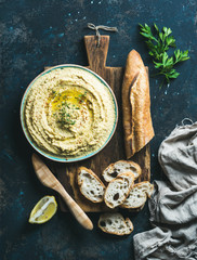 Homemade hummus in blue bowl with lemon, herbs and fresh baguette on rustic wooden serving board...