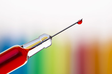 Macro of a single drop of blood hanging at the end of a syringe needle over blurred rainbow background. Shallow DOF.