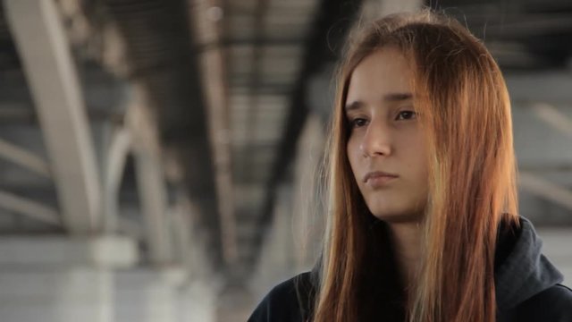 A teenager girl in a hoody with the loose multicolored hair looking into the distance thoughtfully under the bridge. Turning her head aside.