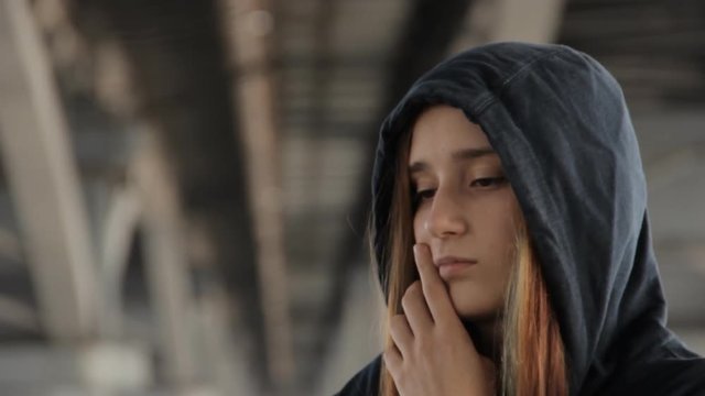 A close up of a teenage girl in a hoody with the hood on and loose multicolored hair spread in the wind under the unfocused bridge pillars scratching a cheek.