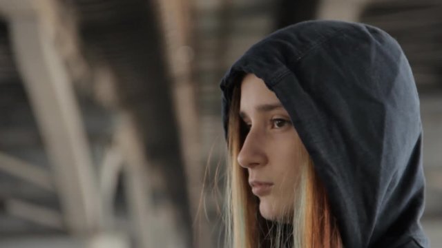 A close up of a teenage girl in a hoody with the hood on and loose multicolored hair spread in the wind under the bridge pillars.