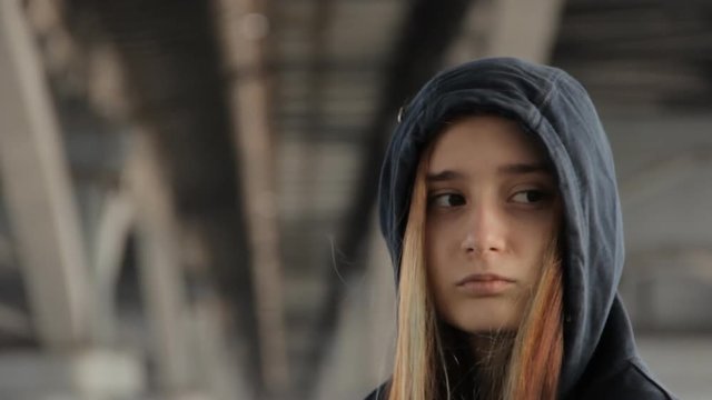 A close up of a teenage girl in a hoody with the hood on and loose multicolored hair spread in the wind under the unfocused bridge pillars watching the car passing by.