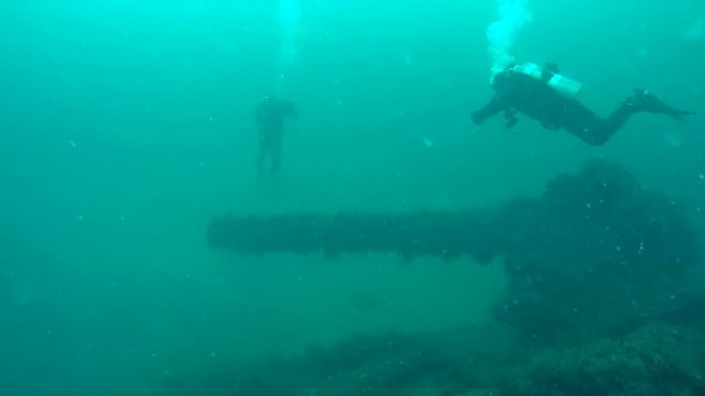 Two divers swim above the cannon on the deck of the sunken warship.
