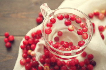 Fresh berries of a forest red cranberry in transparent glass with water among the scattered berries on a white linen cloth on a wooden surface of a table