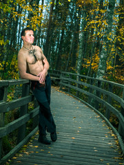 Toned image of a sexy young man with a naked torso which stands near the wooden railing against the backdrop of autumn forest