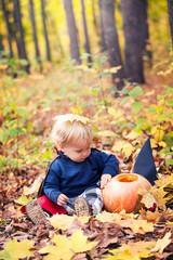 Halloween baby boy in dracula costume (cloak). Child in autumn forest looking at the falling leaves. Halloween pumpkin, witch hat, holiday concept