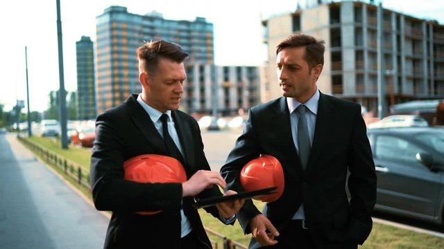 4k. Ultra hd. Two Young successful businessman customers builders in suits walk near newly constructed buildung with orange helmet and Digital Pad. Discussion about investment. Crane and beams BG