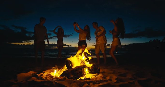 Group of friends dancing around bonfire on the beach at sunset