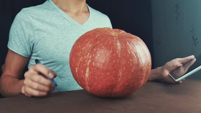 Guy in white shirt use picture on tablet computer as reference and draw on pumpkin jack o lantern 4k