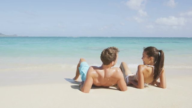 Beach sun tan couple on summer vacation holiday Vacations suntan concept - adults lying down tanning on luxury exclusive paradise beach. Asian woman, Cacuasian man. RED EPIC SLOW MOTION 96 FPS.