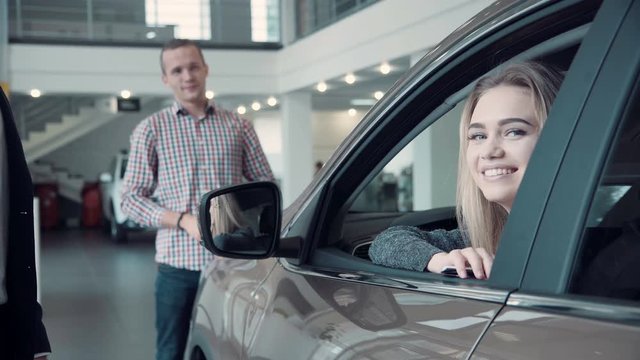 Unrecognizable Sales Manager Gives the Client the Keys from the Car. Then smiling woman customer look straight at the camera, while her husband standing in the background