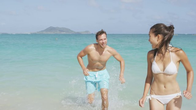 Happy couple having fun on beach running out of water laughing during summer vacation holiday. Multiracial beautiful couple on honeymoon the sun. RED EPIC SLOW MOTION, 96 FPS.