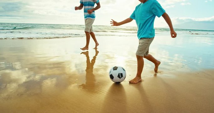 Father and son having fun on the beach at sunset, kicking soccer ball in slow motion