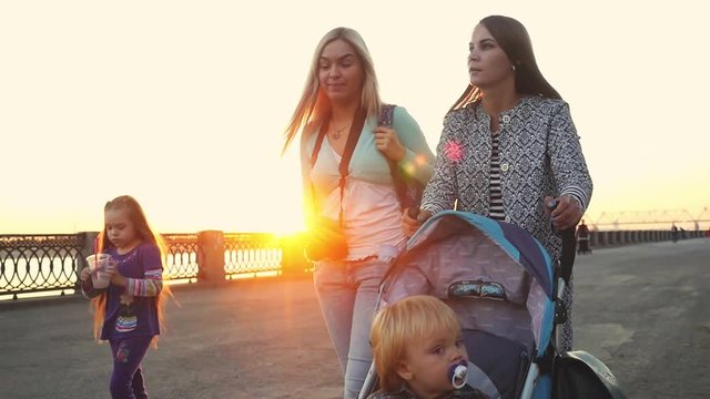 Young beautiful mothers walking together with kids in prams on promenade during sunset in slowmotion. 1920x1080