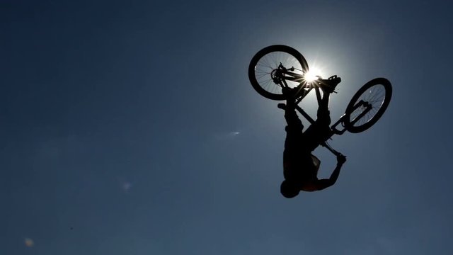 An extreme riders are making free style jumps from a ramp. The young boy with his bicycle is seen as a silhouette in front of the sun.