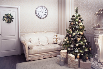 Cozy living room in holiday time. Christmas tree with cones, balls, Garland standing near fireplace, lot of presents under it. On the one wall hangs a needles wreath on the other is watches over sofa